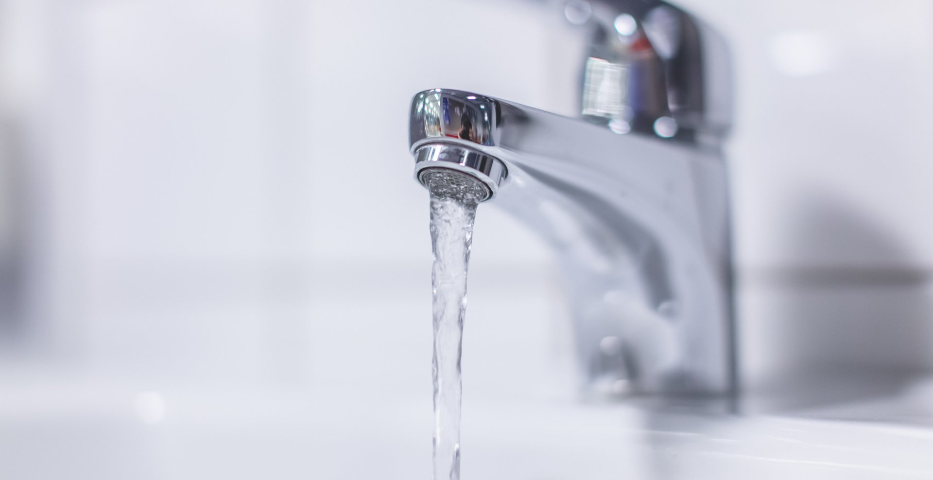 A chrome bathroom faucet flowing with water against a blurred white tiled background.