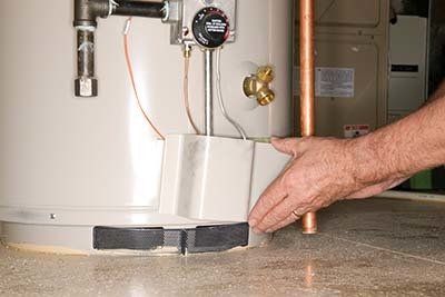A person's hand near the base of a water heater; a drain pan and copper pipes are also visible.