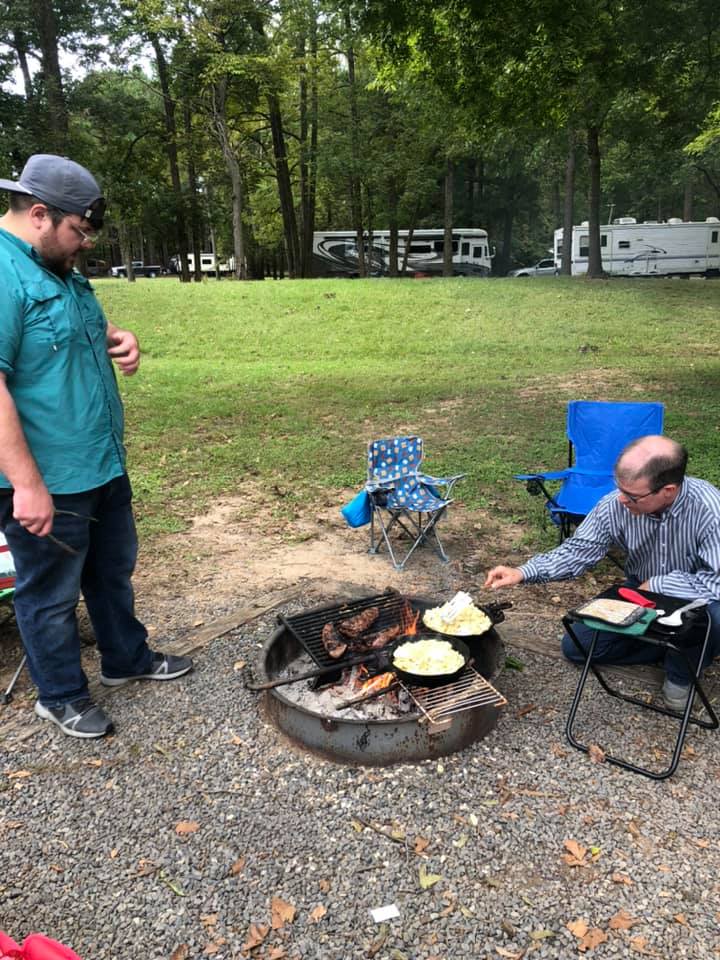 Two men are cooking food over a fire pit at a campground.