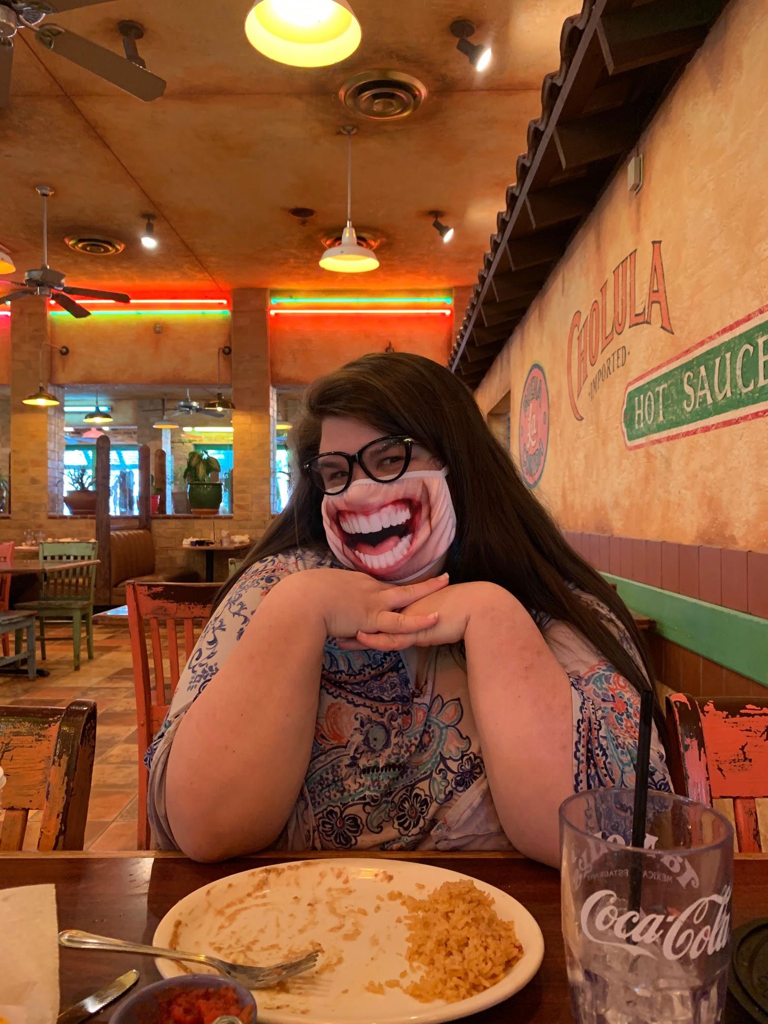 A woman wearing a mask is sitting at a table in a restaurant.