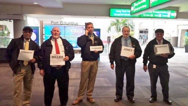 Five men in jackets holding signs with their names in an airport terminal.