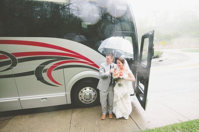Bride and groom exit a bus under an umbrella in the rain. Bus is silver with red and black accents.