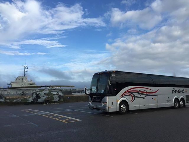 Silver bus parked in a lot, with a cloudy sky and water with a large structure in the background.