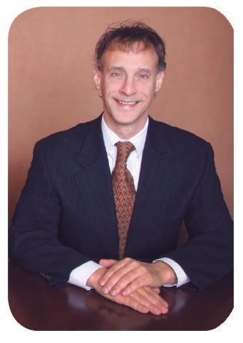 Man in suit, seated at desk, smiling with folded hands. Brown tie, neutral background.