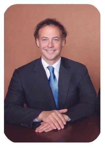 Man in suit, smiling, with arms crossed on table. Blue tie, dark hair, brown background.