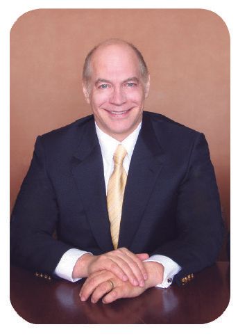 Man in suit at desk, smiling. Leans forward with hands clasped. Light-colored tie.