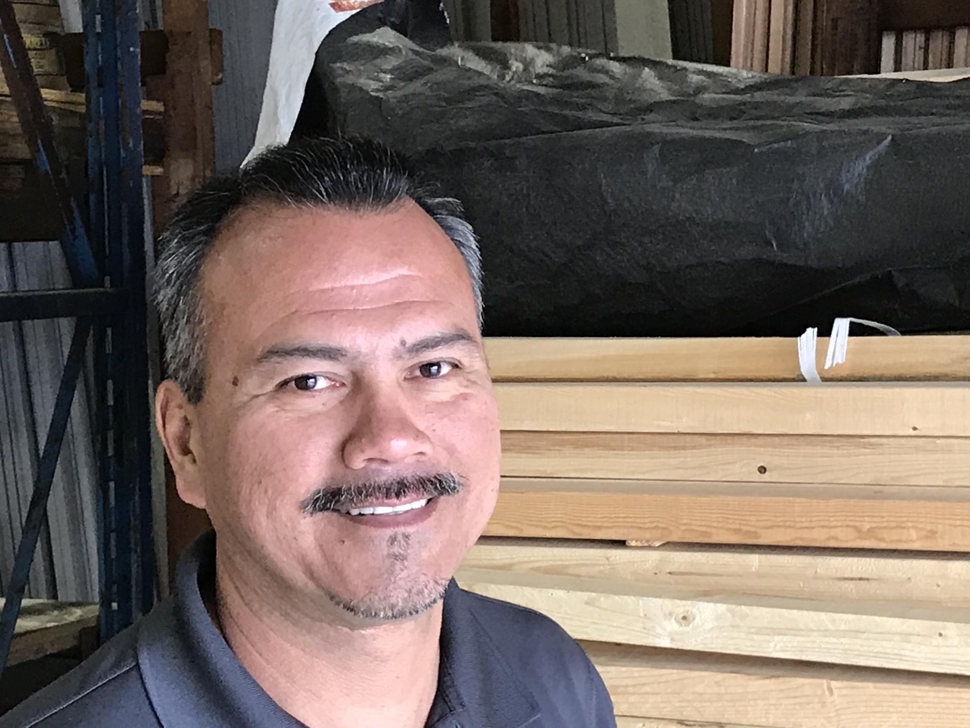 Man with a mustache smiles, standing in a lumber storage area with wood stacks.