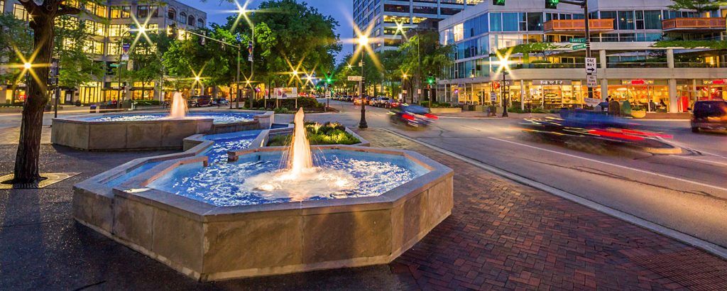 A fountain in a park with cars driving by at night.