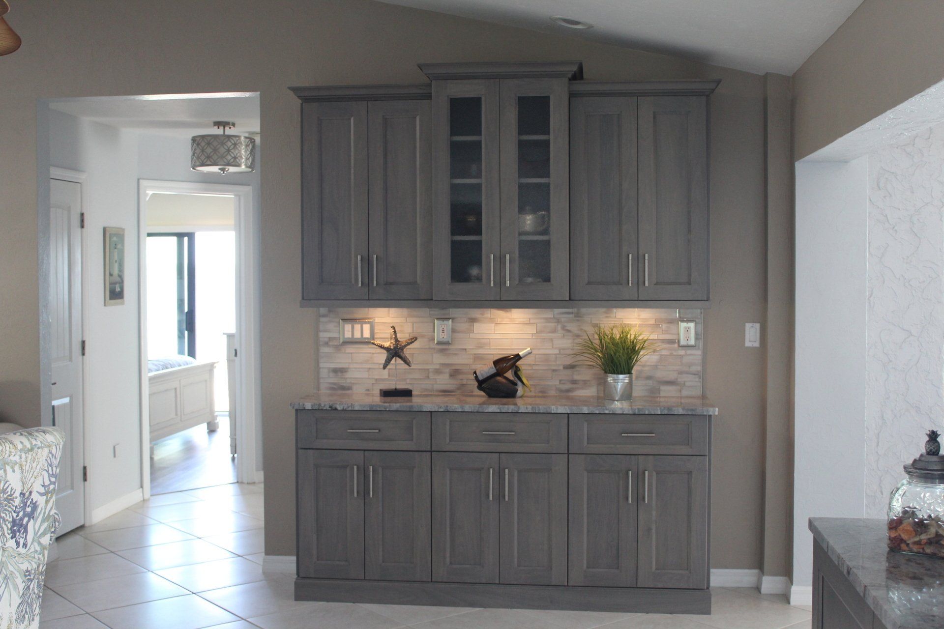 Neutral grey stained dry bar with glass door inserts and crown molding. Accented tile backsplash.