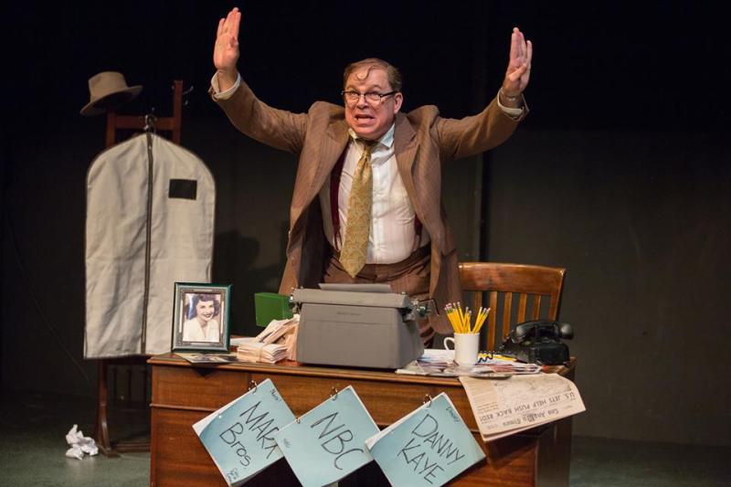 Bernie standing behind his desk holding  both hands in the air.
