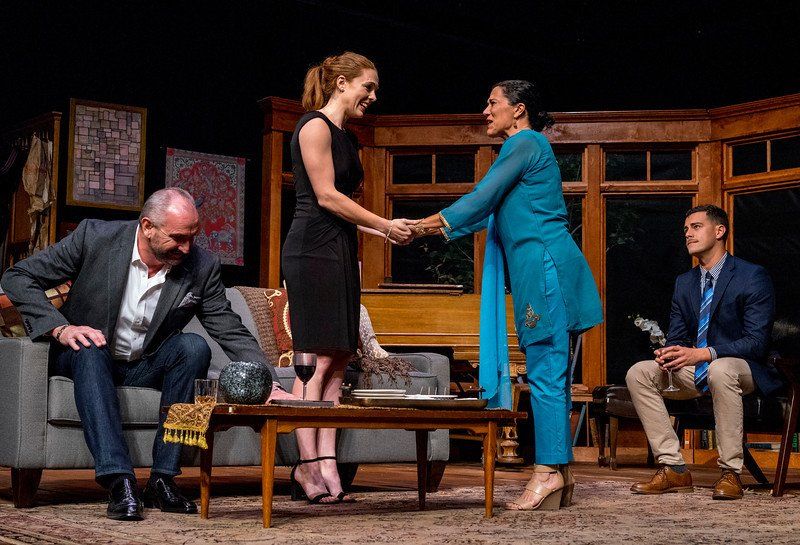 2 Men look on as woman in black dress and woman in blue hold hands in happy greeting