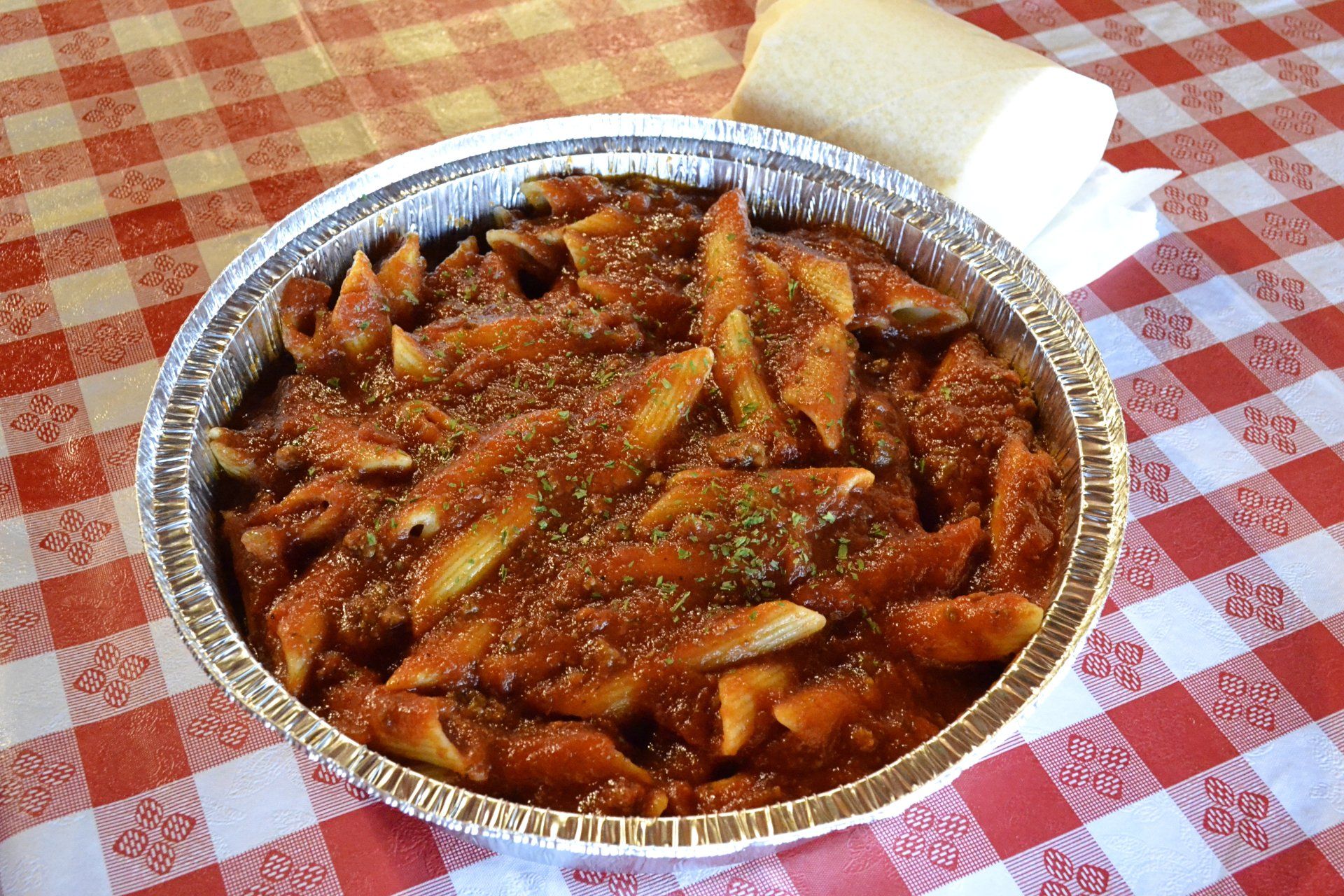 A tin foil container filled with pasta and sauce on a checkered table cloth.