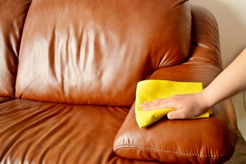 Hand wiping a brown leather couch with a yellow cloth.