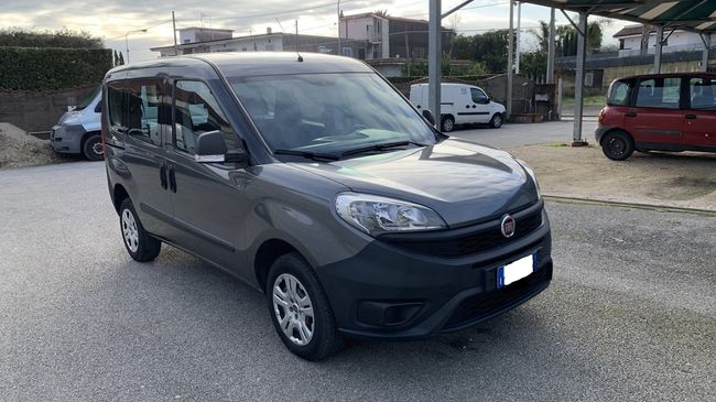 Un furgoncino grigio compatto parcheggiato su un piazzale ghiaioso accanto ad altri veicoli, sotto un cielo nuvoloso.