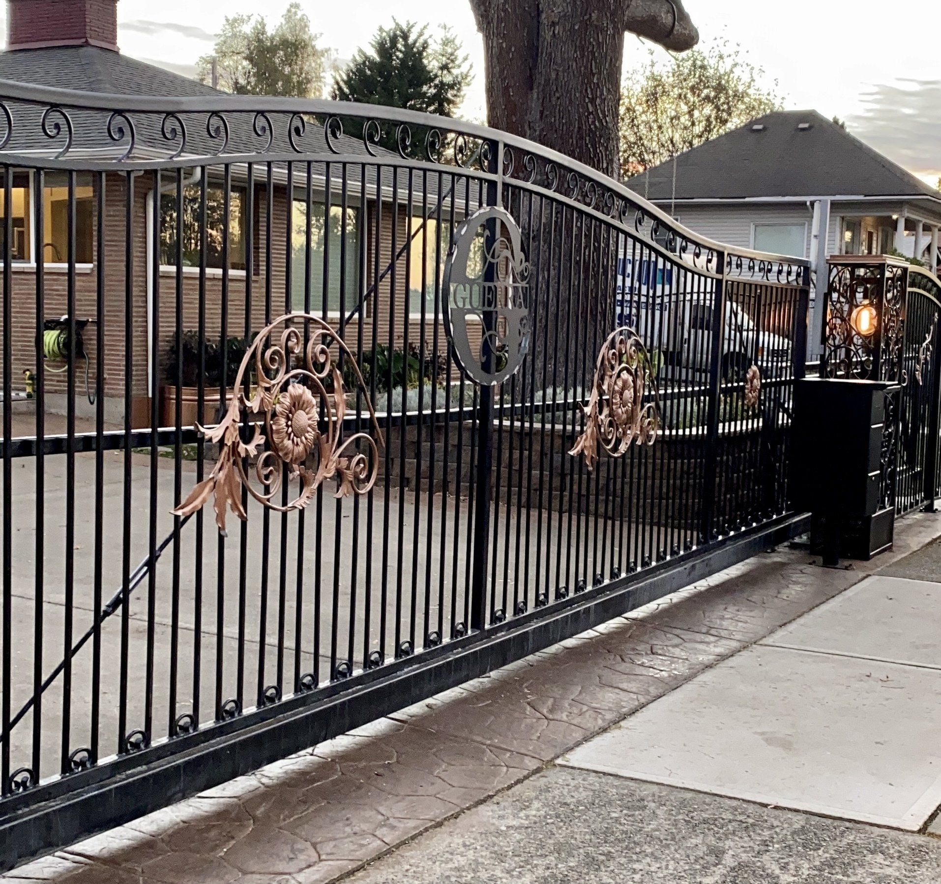 Ornate black metal driveway gate beside a sidewalk, with a lit lamp post and house in the background