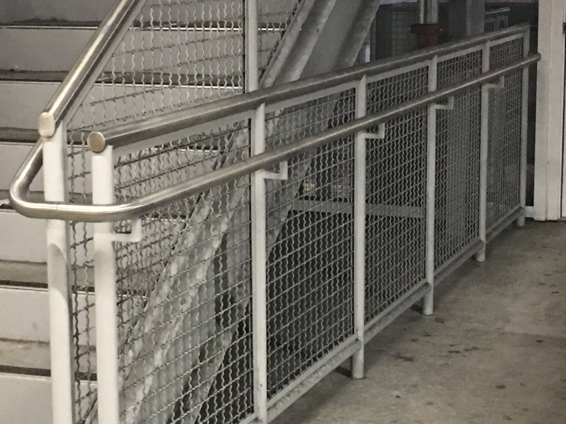 Metal stairway with white handrail beside a grated walkway and wire-mesh safety railing indoors