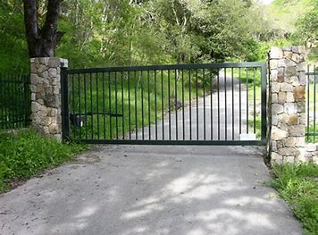 Black metal gate between stone pillars across a paved driveway, with trees and greenery behind it