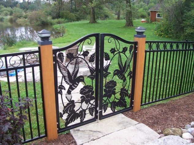 Decorative black metal garden gate with leaf design between orange posts beside a green yard and pond