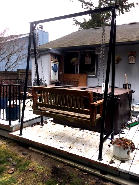 Covered wooden patio with bench and table beside a small house, framed by black metal posts and string lights