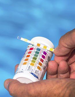 Person testing pool water with a strip and color chart. Blue water background.