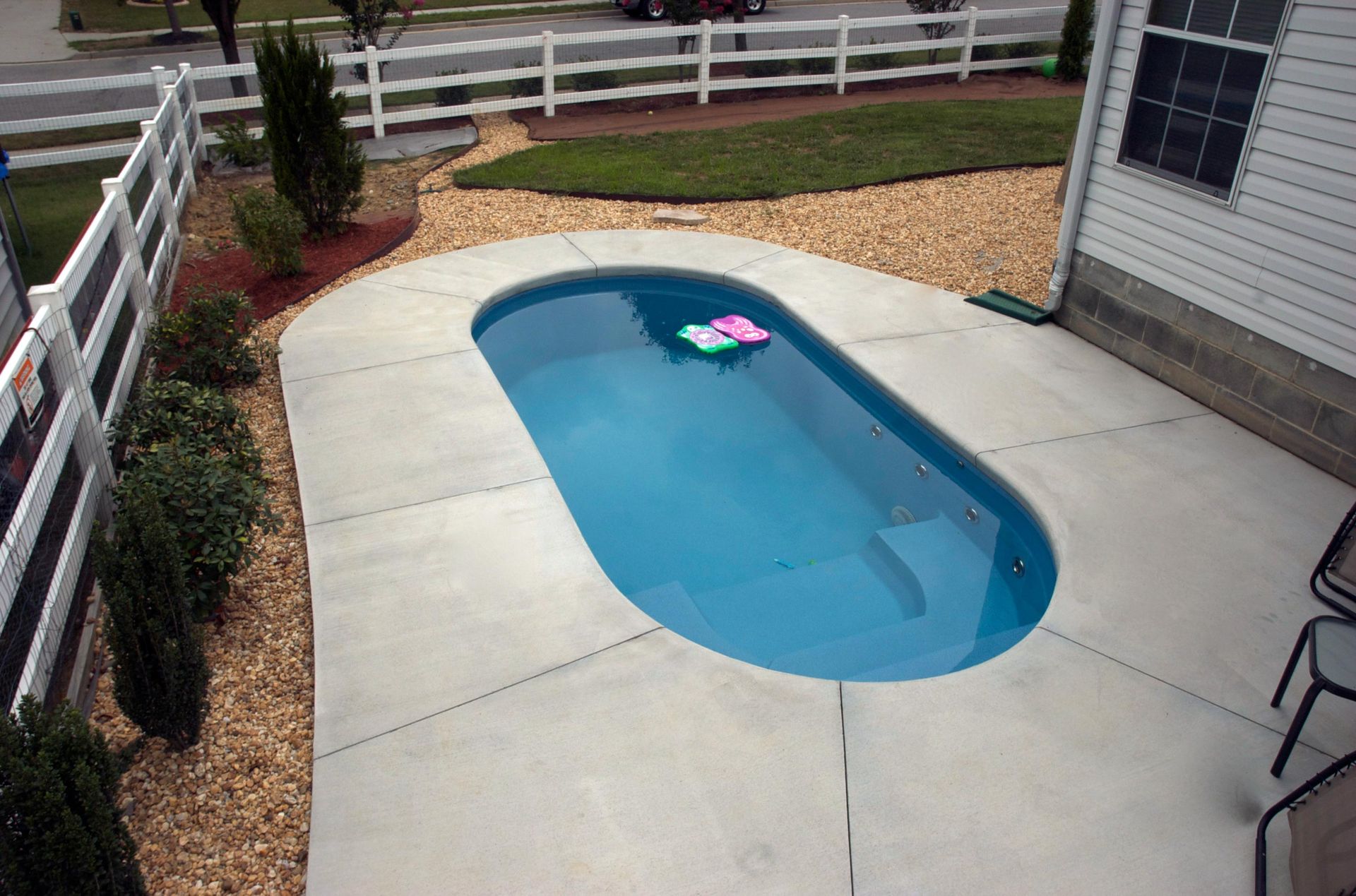 Small oval-shaped pool with blue water and a floaty, surrounded by concrete and landscaping in a backyard.