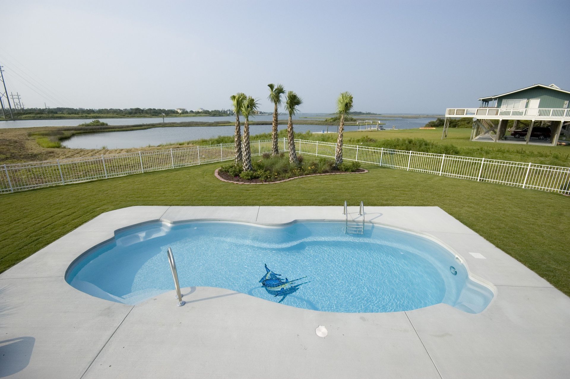 Swimming pool with light blue water, surrounded by concrete, lawn, and palm trees; view of water in the background.