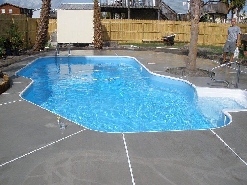 A blue swimming pool with gray concrete edging. A person stands near the pool.