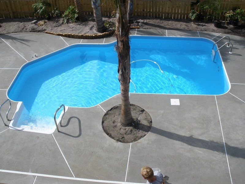 Overhead view of a blue, kidney-shaped pool with a tree in the center; gray concrete patio.