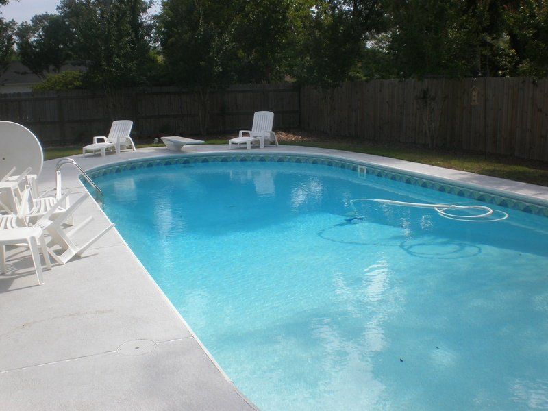 Swimming pool with white lounge chairs on a concrete patio, with a wooden fence and trees in the background.