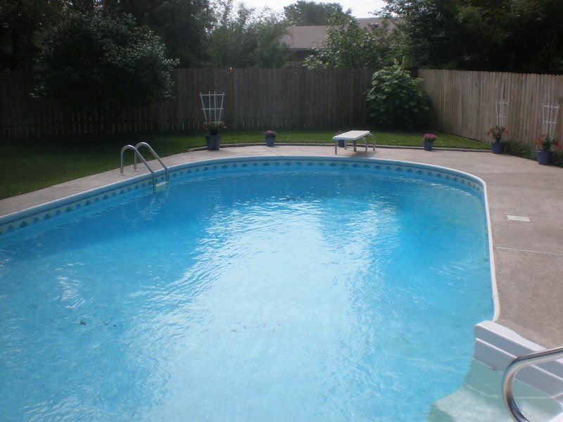 Oval swimming pool in a backyard with a wooden fence and concrete patio.