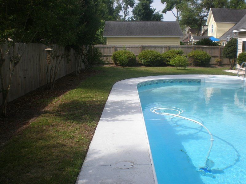 Poolside view with light blue water, concrete edge, green grass, wooden fence, and bushes.