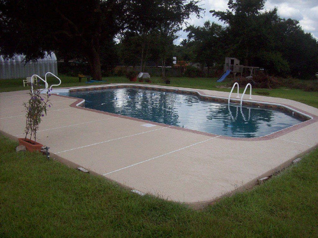 Swimming pool surrounded by concrete, grassy yard, trees in background, cloudy sky.