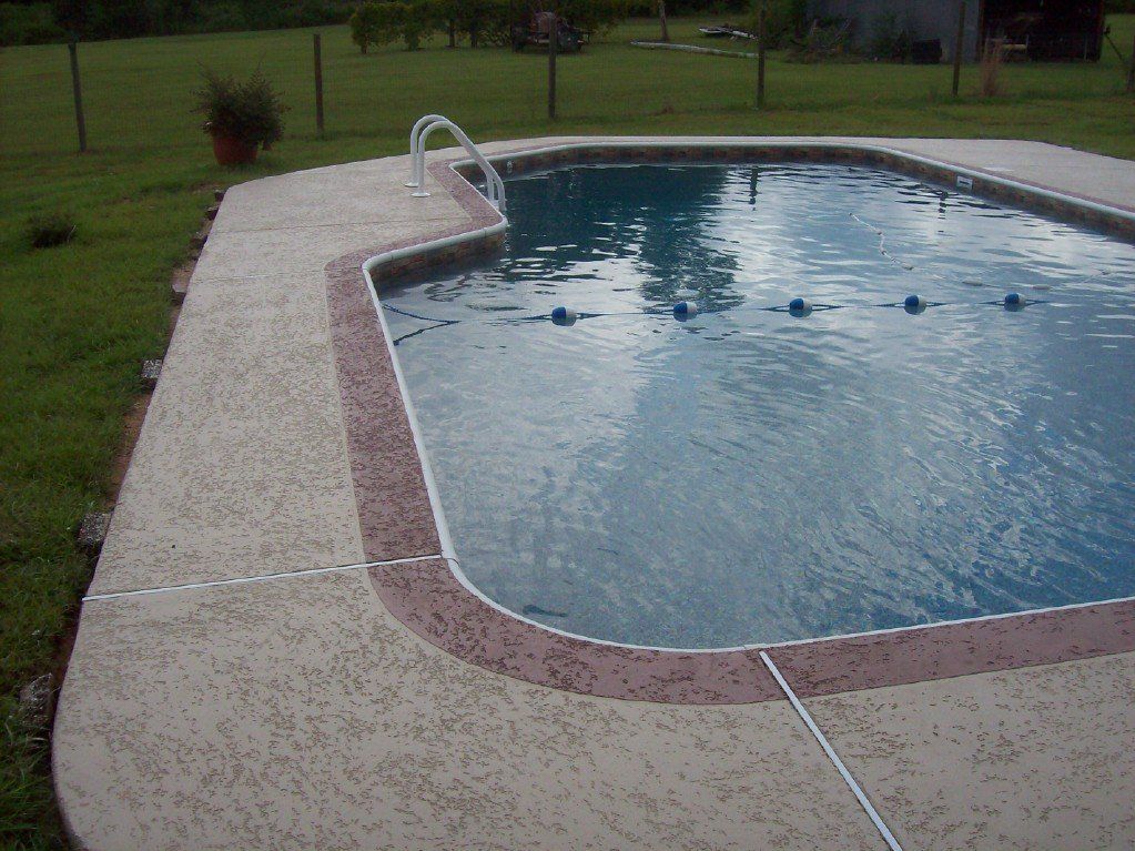 Pool with a concrete deck and brown border, surrounded by grass and a fence.