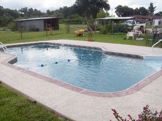 Swimming pool with concrete deck in a backyard, surrounded by grass and trees.