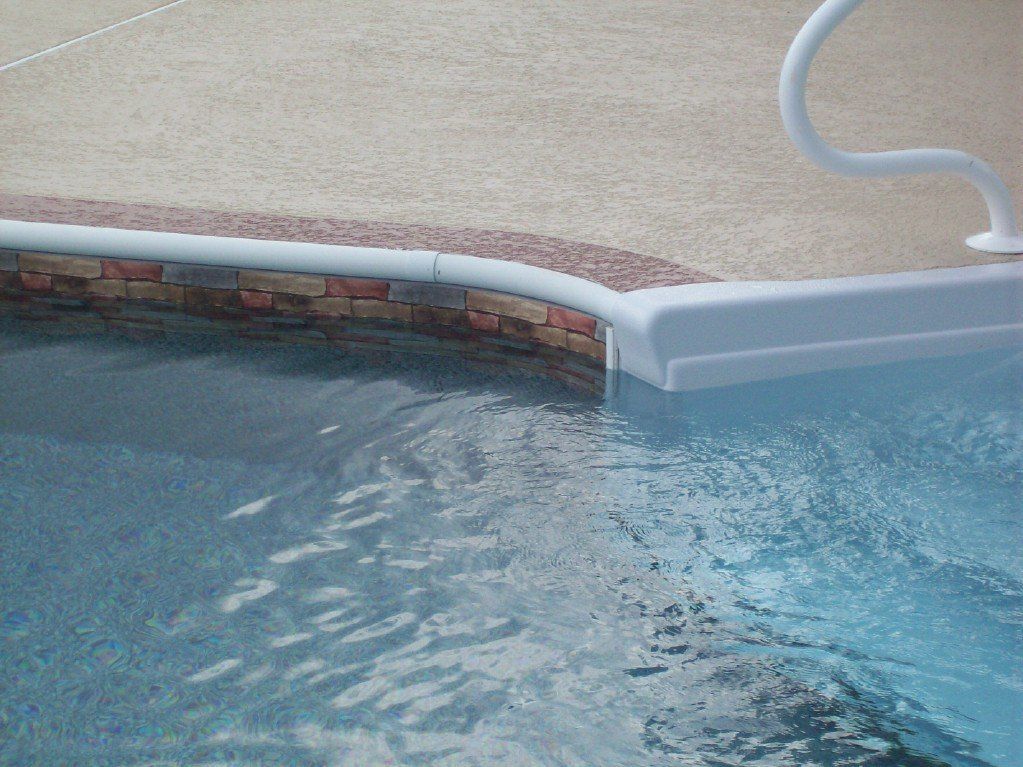 Pool edge with water overflowing into the pool. Light blue water, brick and stone edging, white handrail.