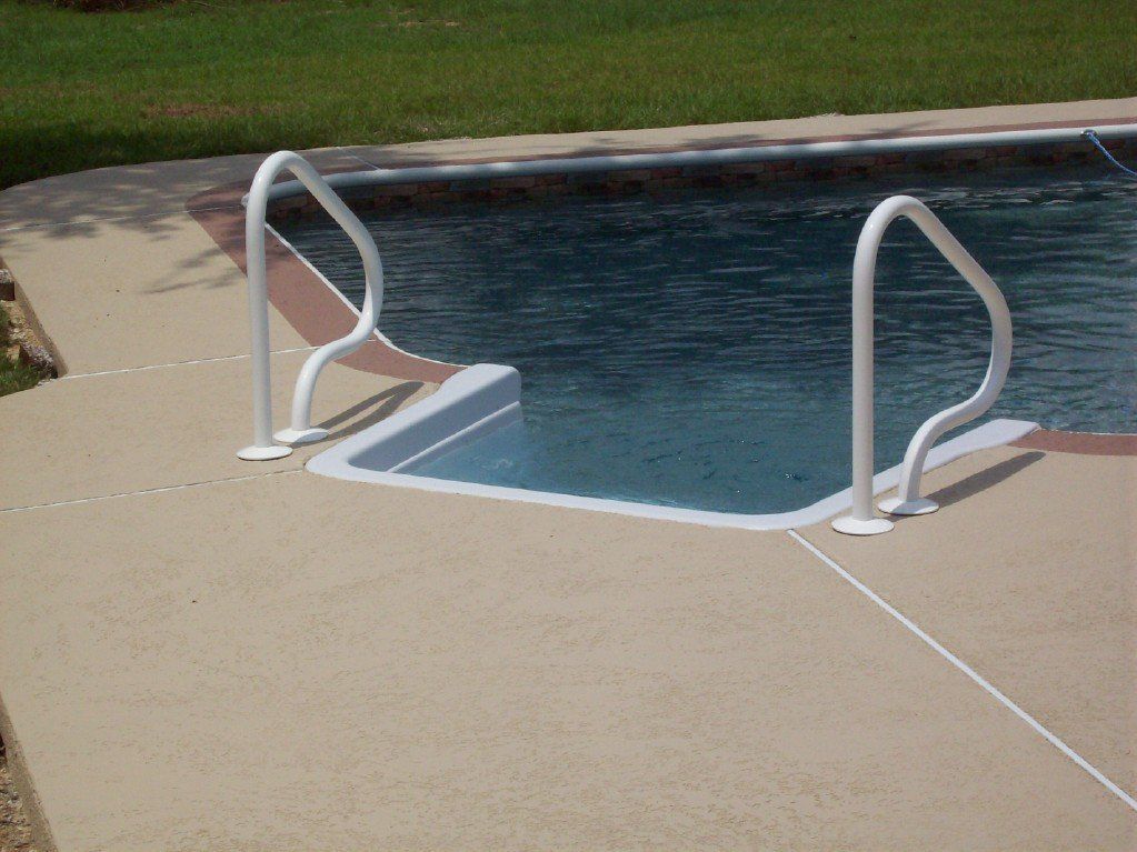 Swimming pool with white handrails and steps, surrounded by light-colored concrete.