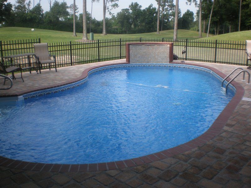 Swimming pool with brick patio, black fence, and view of green lawn and trees.