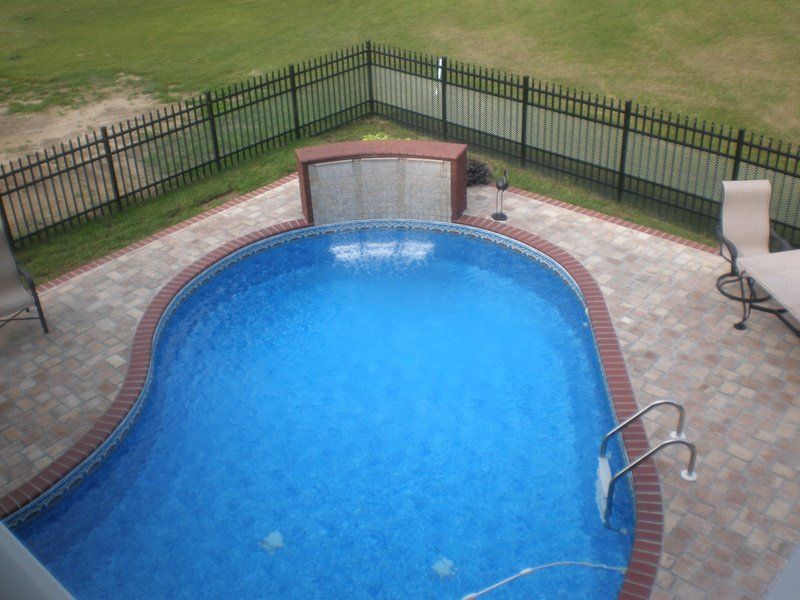 Swimming pool with brick border, water feature, and black fence in backyard.