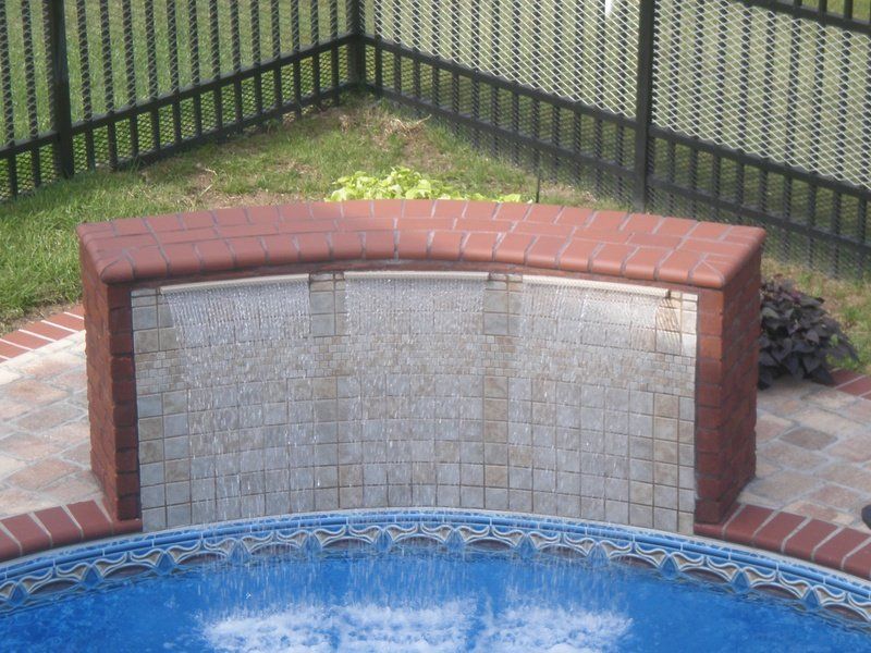 Brick-framed water feature spilling into a blue pool. A black fence and brick patio surround the feature.
