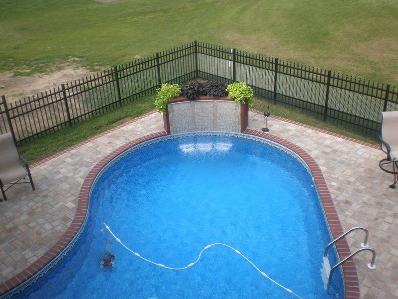 Pool with brick border, black fence, and green lawn. Water feature with plants and blue water.