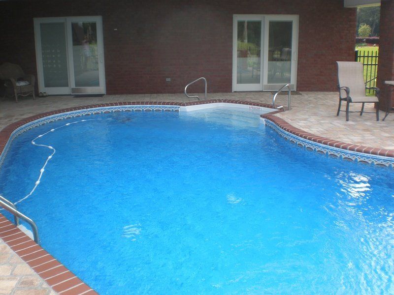 Blue swimming pool with brick trim under a covered patio. White doors and a chair are visible.