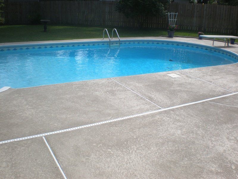 A concrete patio surrounds a blue oval pool with a ladder. A wooden fence and grass are in the background.