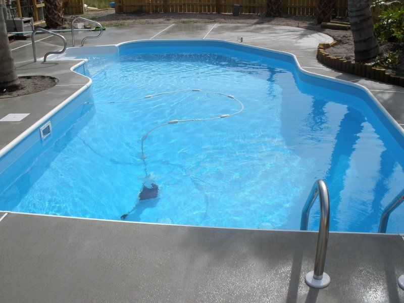 Swimming pool with blue water and gray concrete surrounding it. A robotic cleaner is in the pool.
