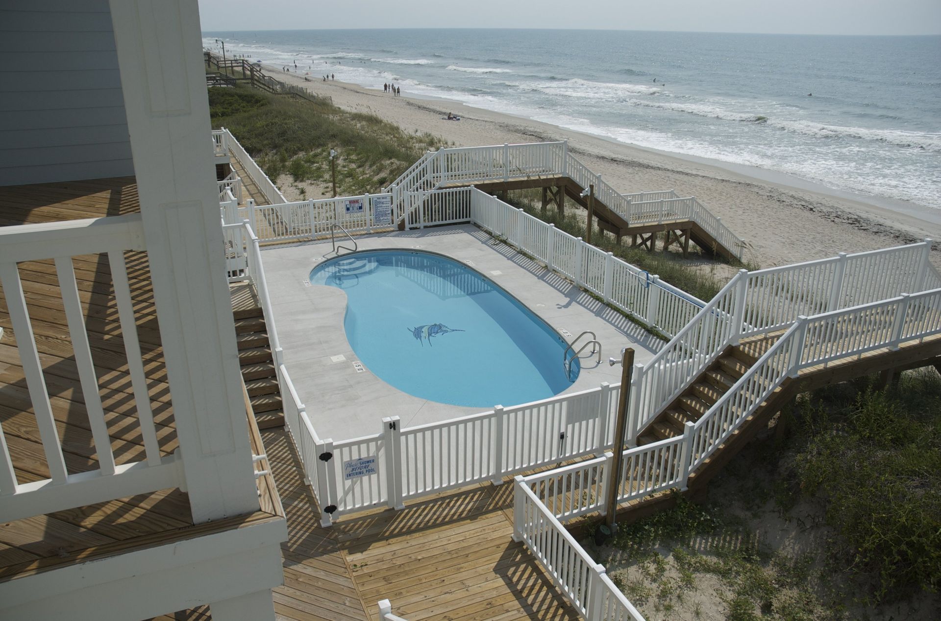 Pool next to beach with stair access. White fencing surrounds the pool, ocean in the background.