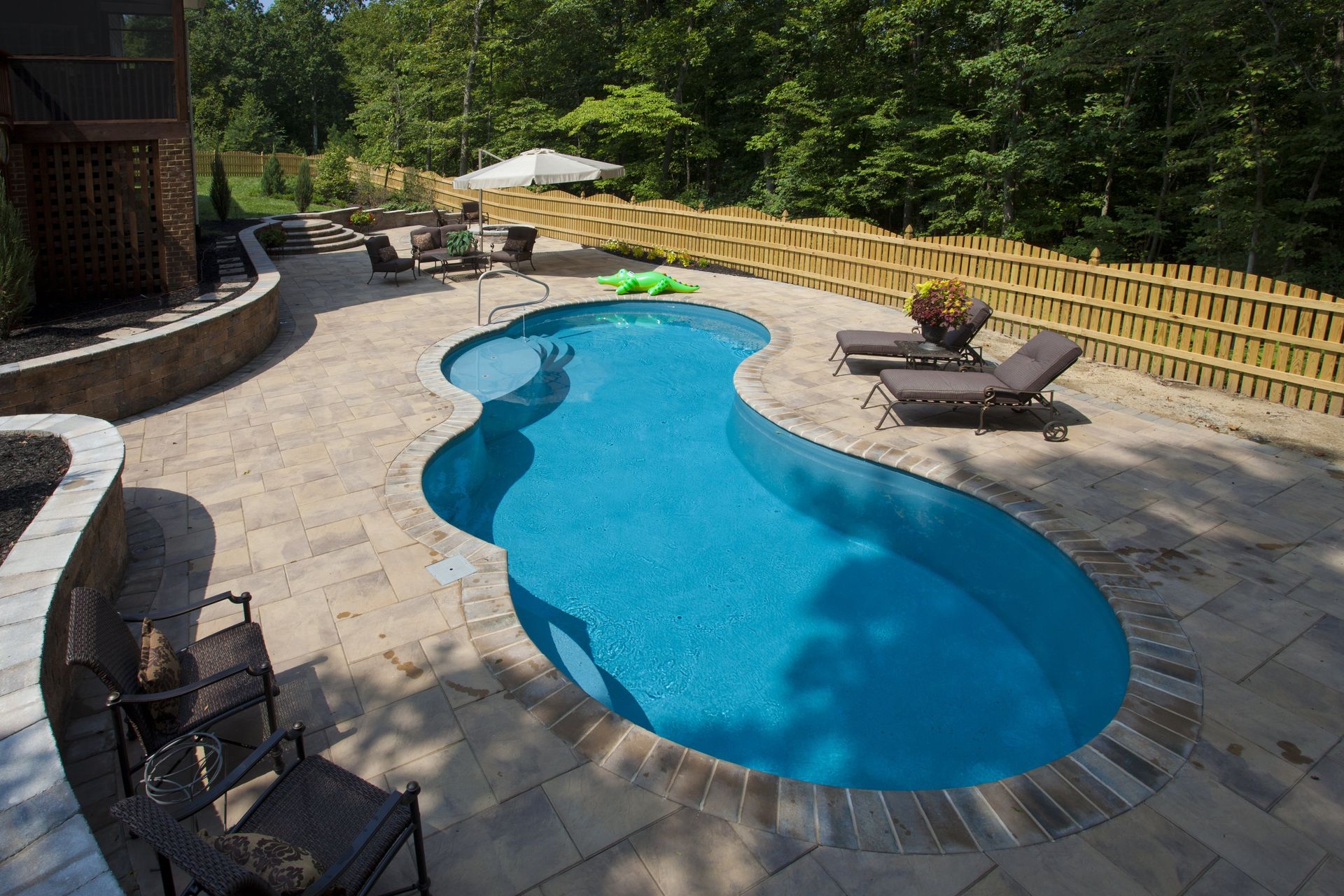 Swimming pool with blue water and surrounding stone patio, lounge chairs, and wooden fence.