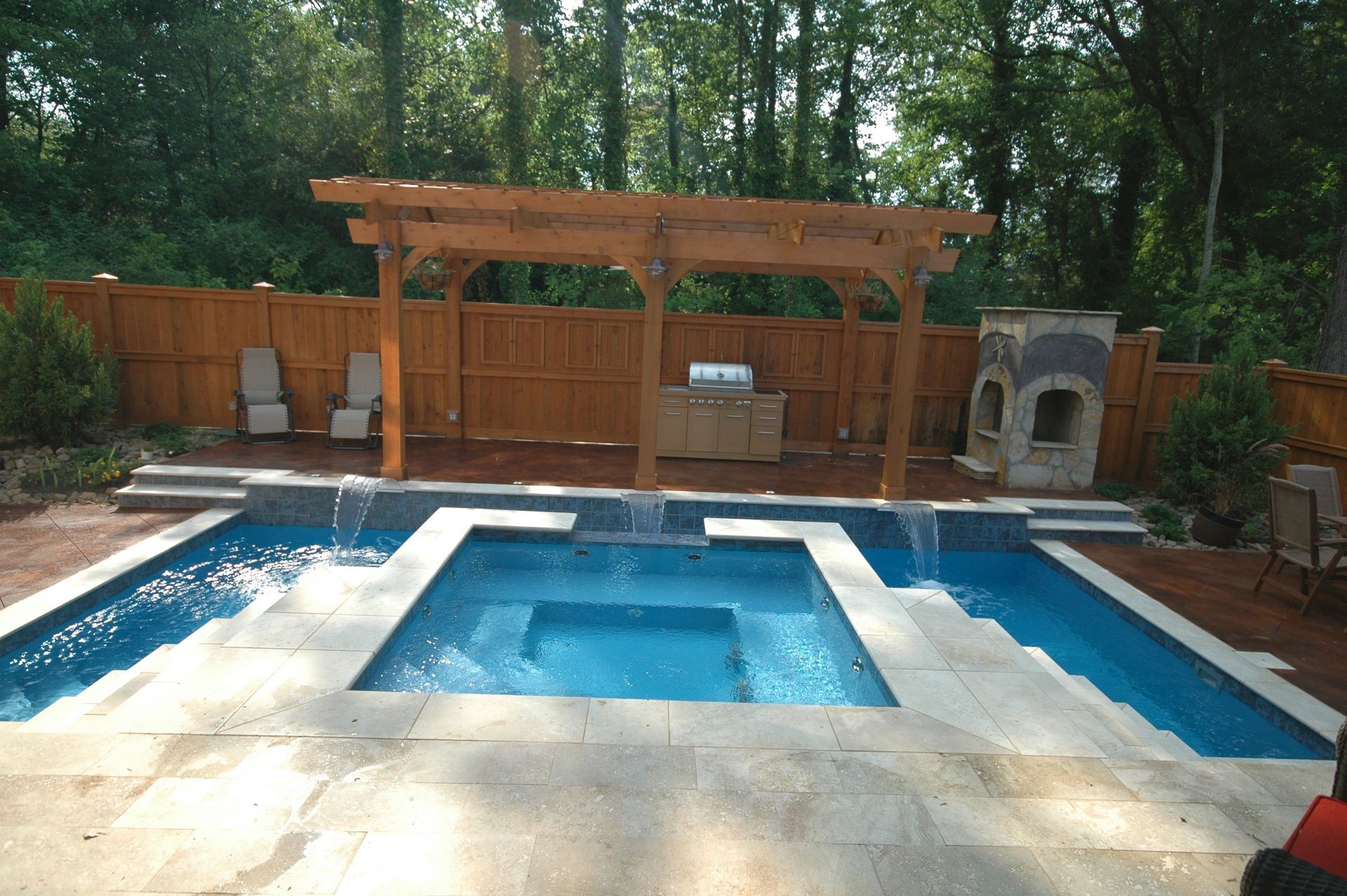 Backyard pool with pergola, grill, and stone fireplace, blue water, and wooden fence.