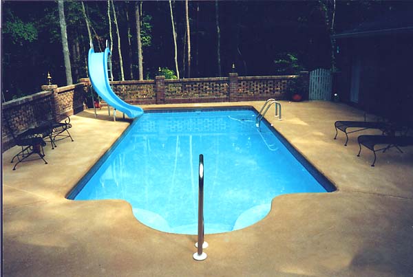 Swimming pool with blue water, a slide, and a brick wall in a wooded area.