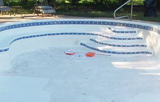 Empty, white in-ground swimming pool with blue tile border and steps. Cleaning tools lie inside.