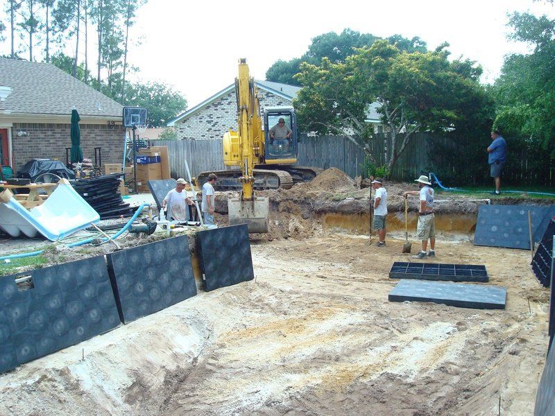 Construction of an in-ground pool; excavator, workers digging, dark wall blocks, sandy ground, backyard.