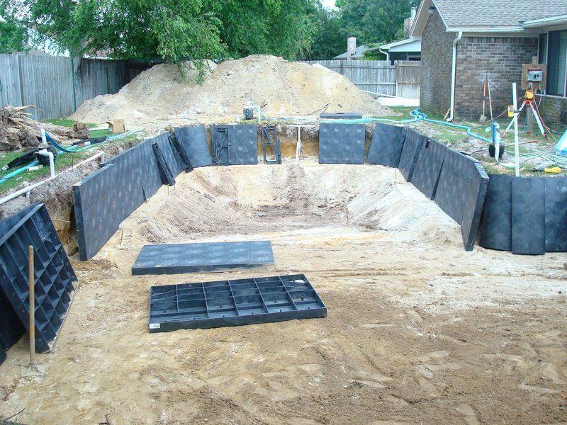 Pool under construction: excavated ground with retaining walls and sand pile in a backyard setting.